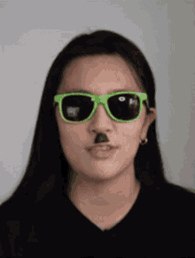 a girl wearing green sunglasses has a fake mustache on her face