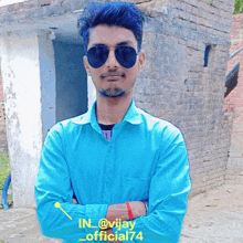 a young man wearing sunglasses and a blue shirt is standing with his arms crossed in front of a brick building