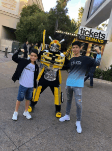 two boys pose with a bumblebee mascot in front of a sign for tickets on demand