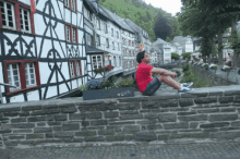 a man in a red shirt sits on a stone wall in front of a row of half timbered houses