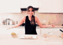 a woman in a black tank top is standing in a kitchen with her arms outstretched and a plate of food on the counter