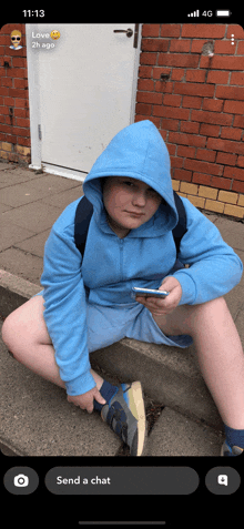 a boy wearing a blue hoodie is sitting on the sidewalk looking at his phone