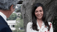 a woman in a floral shirt smiles while standing next to a man in a suit in front of a tree