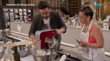 a man is using a red mixer while a woman looks on in a kitchen with #masterchefargentina on the screen