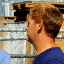 a man in a blue shirt is standing in front of a chain link fence and a stack of wooden pallets