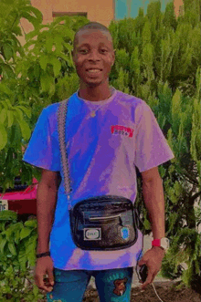 a young man wearing a purple tie dye t-shirt and blue jeans is standing in front of a tree .