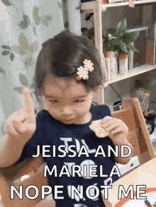 a little girl with a flower in her hair is sitting at a table eating a cracker and giving a thumbs up