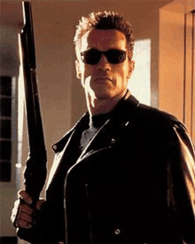 a man wearing sunglasses and a leather jacket is holding a gun .