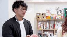 a man wearing glasses and a black jacket stands in front of a shelf full of magazines including one called you