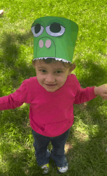 a little girl wearing a pink shirt and a green paper hat with big eyes