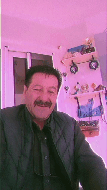 a man with a mustache and glasses is smiling in front of a pink wall with shelves