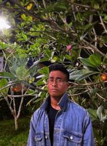 a man wearing glasses and a blue denim jacket is standing in front of a tree .