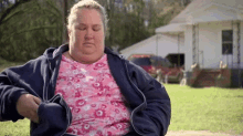 a very fat woman wearing a pink shirt and a blue jacket is sitting in front of a house .