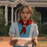 a girl in a blue dress and red scarf is holding a piece of paper with netflix written on it