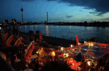 a group of people sitting at tables near a body of water at night