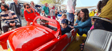 a little boy is sitting in a red toy car with a woman holding his hand