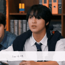 a young man in a white shirt and tie is sitting in front of a bookshelf with the year 2016 written on a piece of paper