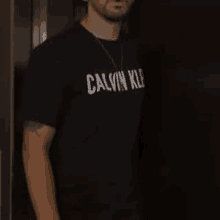 a man wearing a black calvin klein t-shirt and a necklace is standing next to a door .