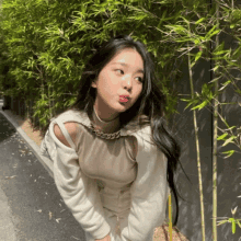 a woman wearing a white sweater and a chain around her neck is standing in front of a row of bamboo trees