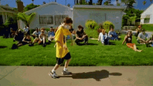 a boy in a yellow shirt is walking on a sidewalk in front of a group of people sitting on the grass