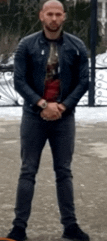 a man in a leather jacket and jeans is standing with his hands folded .