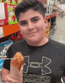 a young man wearing a black under armour shirt is holding a fried chicken wing in a paper bag .