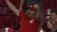 a man in a red shirt and hat is raising his arms in the air while sitting in a stadium .