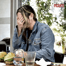 a man in a denim jacket sits at a table with a glass of water and a sandwich on it