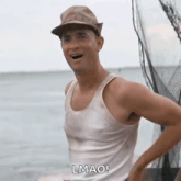 a man wearing a hat and a white tank top is standing on a boat in the ocean .
