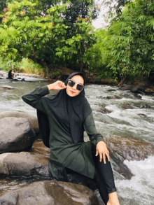 a woman wearing a hijab and sunglasses sits on a rock near a river