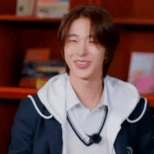 a young man in a school uniform is smiling while sitting in front of a bookshelf in a library .