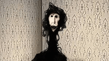 a black and white photo of a puppet with a long haired head standing in front of a wall with a patterned wallpaper .