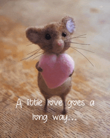 a mouse holding a pink heart with the words a little love gives a long way below it