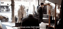 a man is sitting in front of a mirror in a suit shop talking to another man .