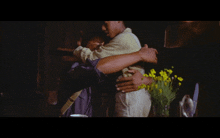 two men hugging in a dark room with a vase of yellow flowers in the background