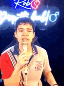 a young man is singing into a microphone in front of a neon sign that says ' kobe ' on it