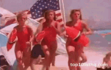 a group of women in red swimsuits are running on a beach with an american flag in the background .