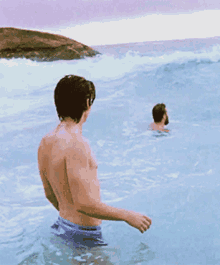 a shirtless man is standing in the ocean watching another man swim