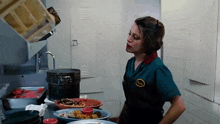 a woman in a green uniform is standing in a kitchen with plates of food and a pot .