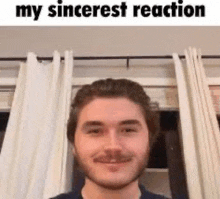 a man with a beard is smiling in front of a window with curtains behind him .