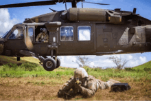 a military helicopter is taking off from a grassy field with a soldier laying on the ground