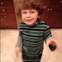 a young boy in a green and black striped shirt is standing in a kitchen holding a remote control .