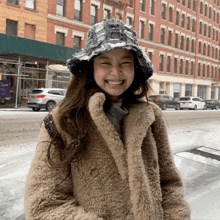 a woman wearing a hat and a fur coat is smiling in front of a snowy street .