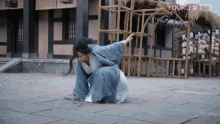 a man in a blue robe is kneeling down on the ground in front of a building that says youku on the bottom