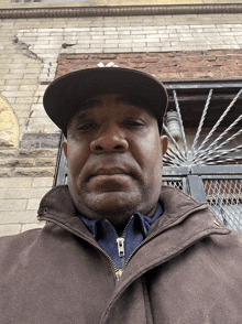 a man wearing a hat and a brown jacket stands in front of a brick building