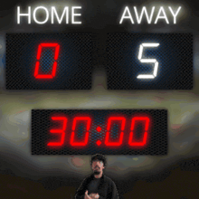 a man stands in front of a scoreboard which says home and away