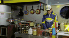 a man in a yellow and blue jacket is standing in a kitchen with pots and pans hanging on the wall