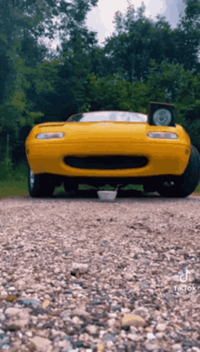 a yellow sports car is parked on a gravel road with a bowl on the ground in front of it .