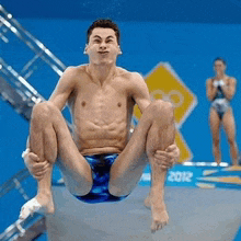 a shirtless man is sitting on a diving board with a 2012 sign in the background