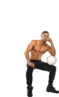 a shirtless man sits with his hand on his chin while holding a white volleyball